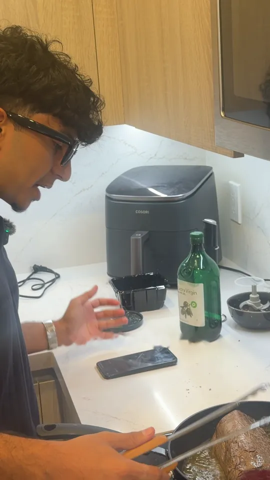 Malik, wearing Meta glasses, turns a beef fillet in a hot pan with tongs. His phone lies on the counter running Glutt.